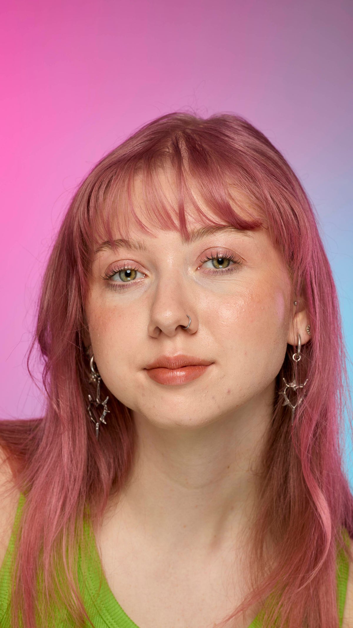 The image shows a fair-skinned model on a pink and blue gradient background. They have pink hair and subtle shimmering lips from using the Crackled Copper lip cream.